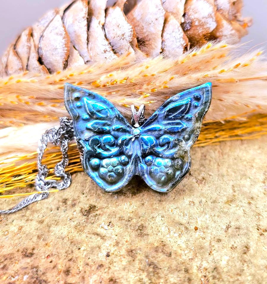 A butterfly-shaped pendant necklace with iridescent blue and green enamel-like detailing, resting on a textured stone surface with dried grass and a pinecone in the background.