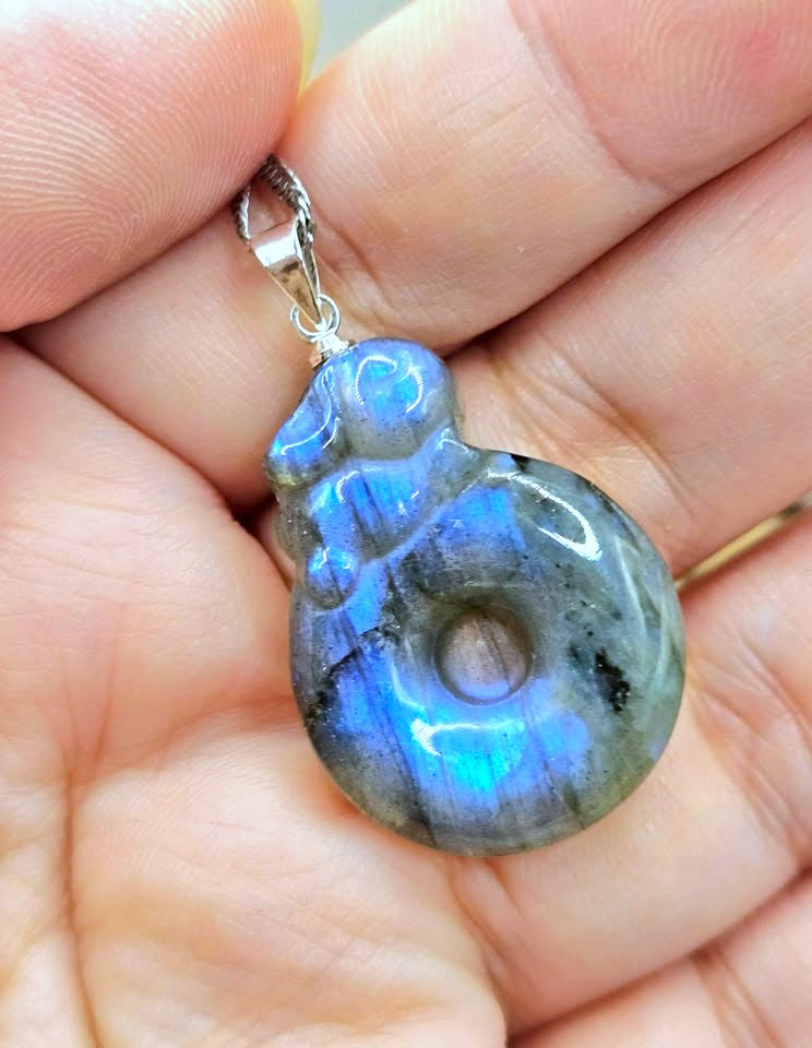 A polished labradorite pendant with vibrant blue flashes and gray-green base tones, held in a person's hand. The pendant is carved with a stylized figure and hangs from a silver bail.