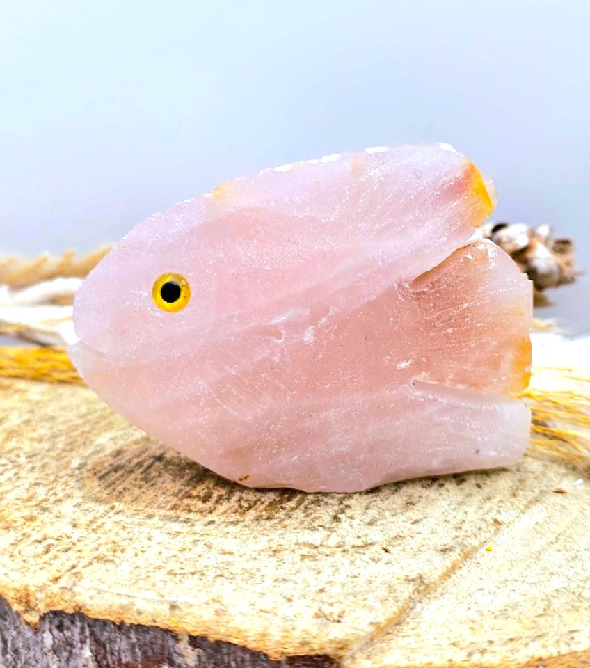A pink rose quartz crystal fish pendant or charm with a yellow-rimmed eye, resting on a textured wooden slice with a blurred light blue background.