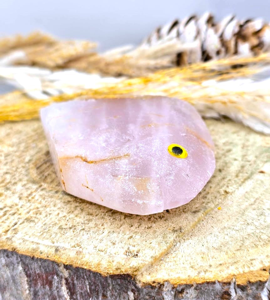 A pale pink crystal stone with a small yellow and black eye detail, resting on a rustic wood slice with dried grass and a pine cone in the background.