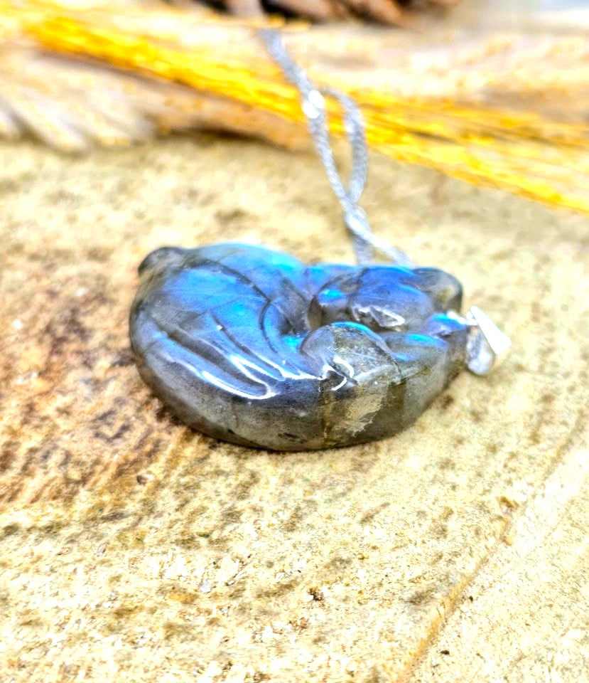 A polished labradorite pendant necklace with a silver-toned bail and chain, resting on a textured stone surface with dried grass in the background.