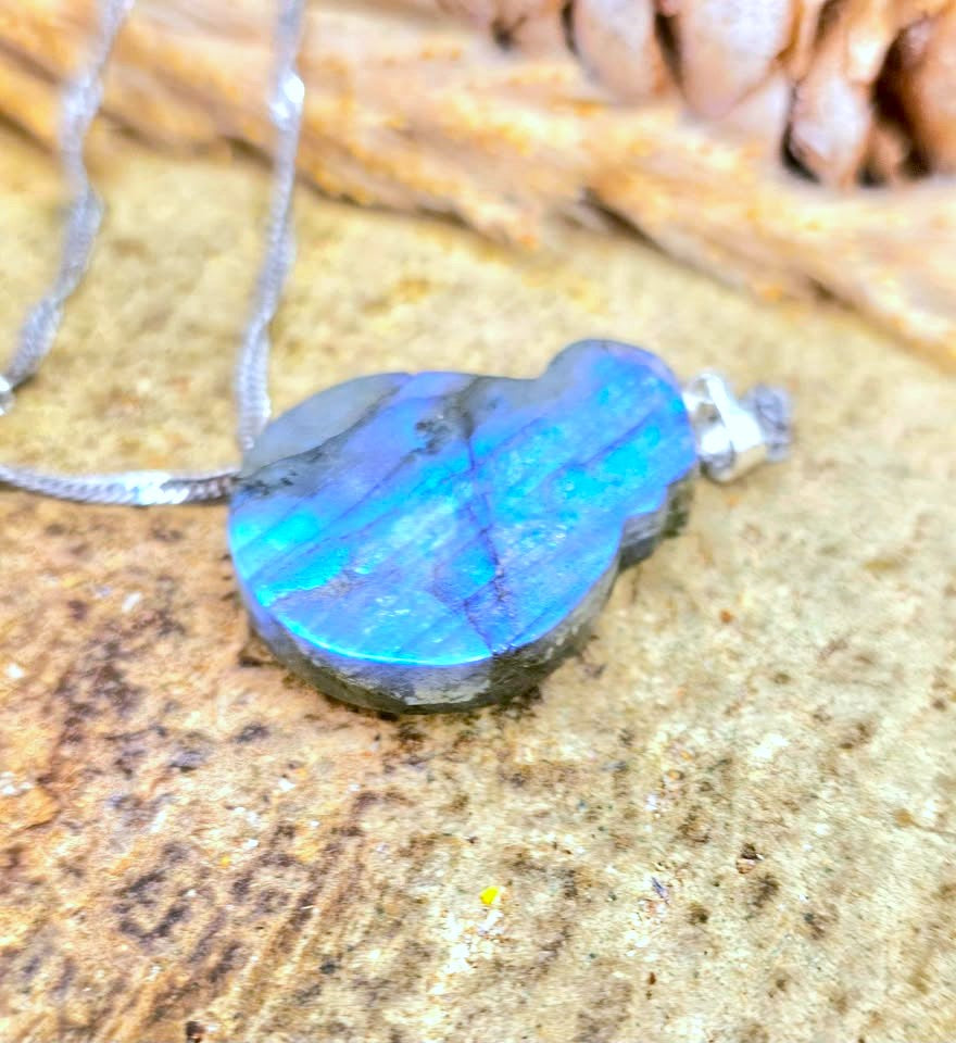 A close-up of a labradorite pendant necklace resting on a textured stone surface. The pendant is highly iridescent, showing vibrant blue, teal, and green flashes, set in a silver-toned metal bezel with a silver chain.