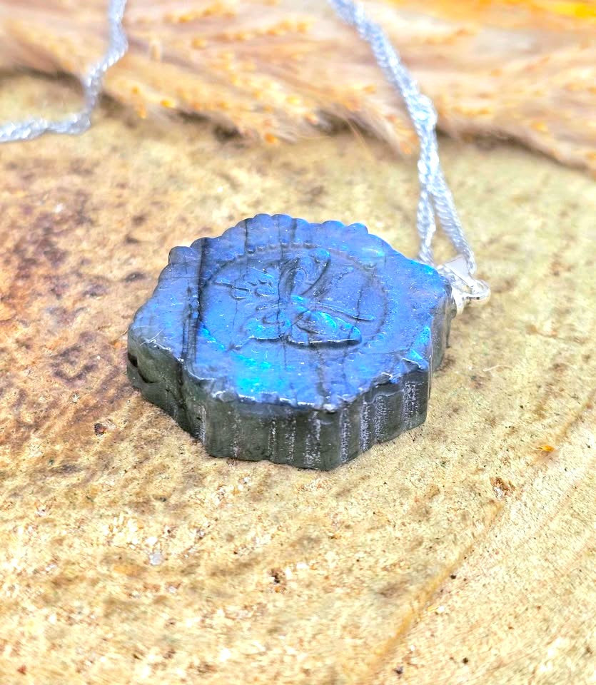 A dark gray, irregularly shaped pendant with a blue iridescent top and an engraved symbol, hanging from a silver chain on a textured sandy surface.