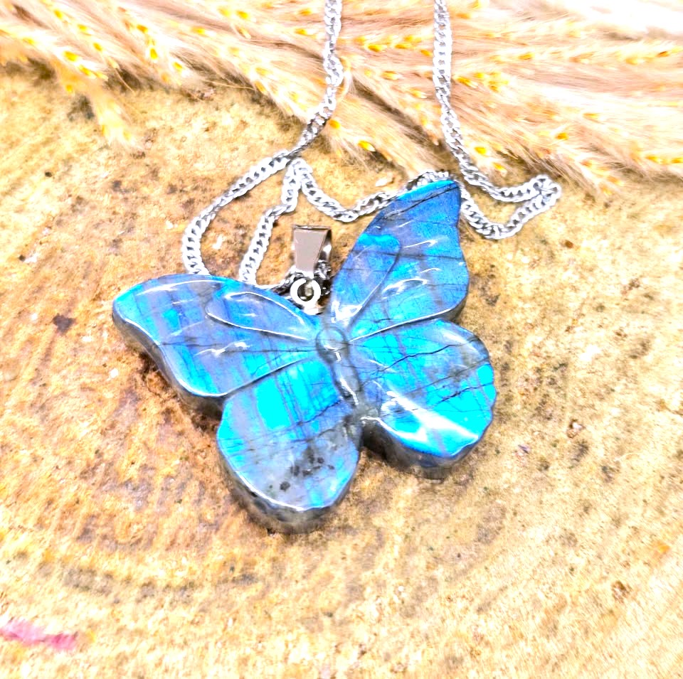 A butterfly-shaped pendant necklace made from iridescent blue labradorite stone with a silver chain, displayed on a textured wooden surface with dried grass.