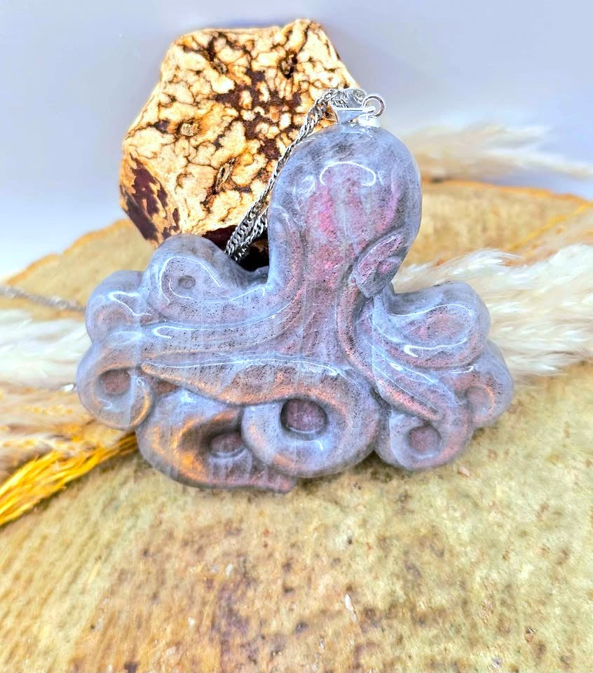 A carved octopus-shaped pendant necklace with a textured, weathered finish in muted grayish purple and pink tones, resting on a natural cork and dried grass background.
