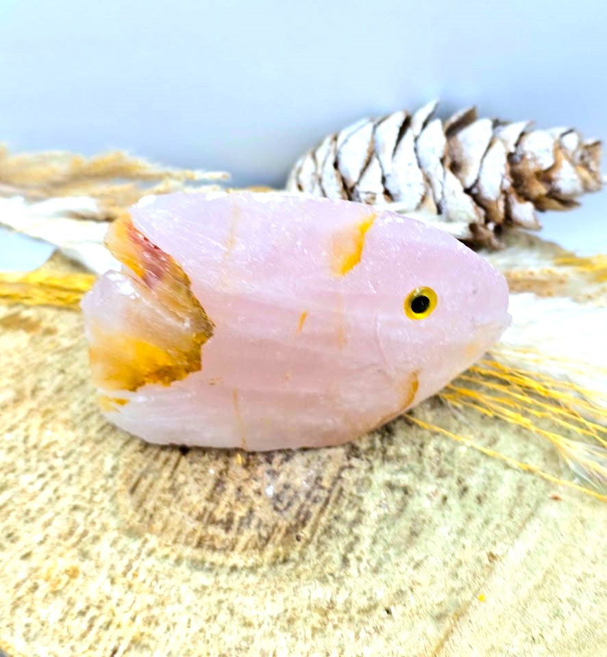 A pale pink crystal fish figurine with yellow-orange accents and a small yellow eye, displayed on a textured beige surface with a pinecone and dried grass in the background.