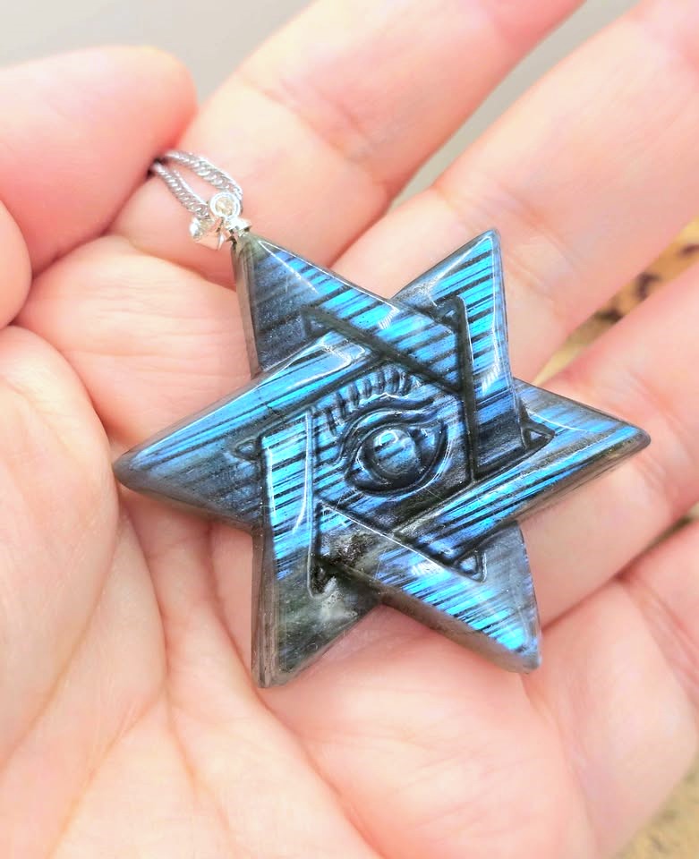 A close-up of a hand holding a six-pointed star pendant with a blue and black striped pattern and an eye design in the center, attached to a silver chain.