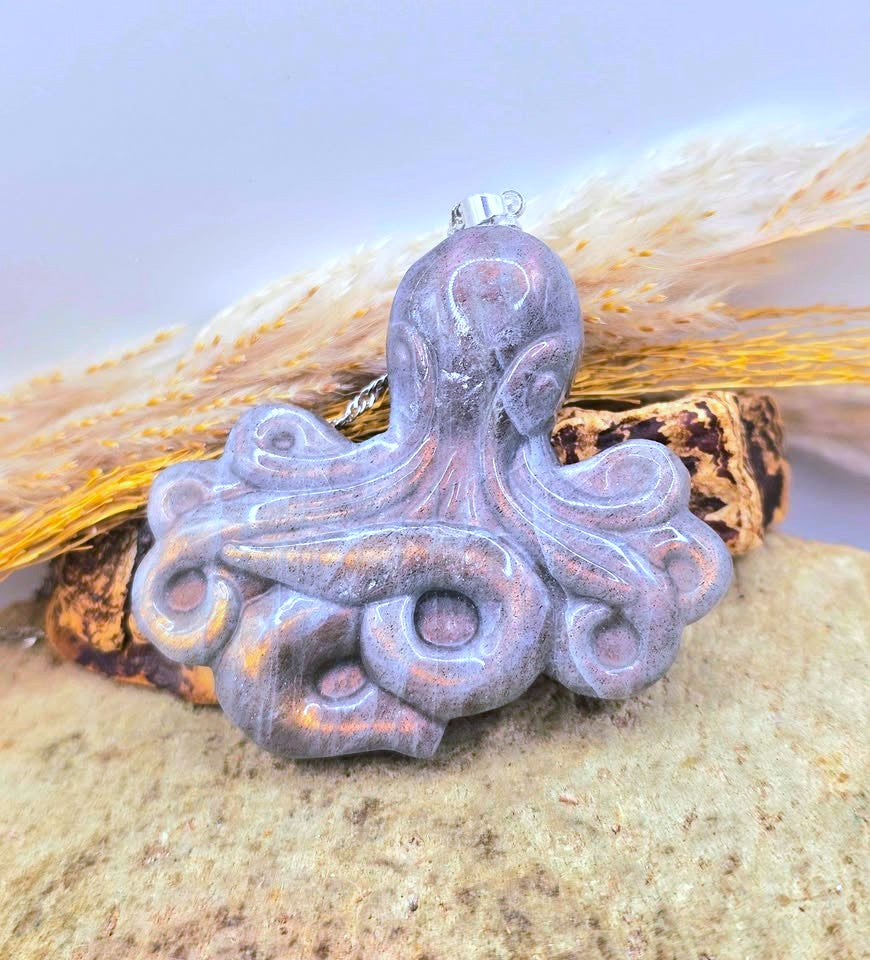 A carved octopus pendant with a distressed lavender-purple finish and subtle coppery highlights, resting on a piece of driftwood with dried grass against a light background.