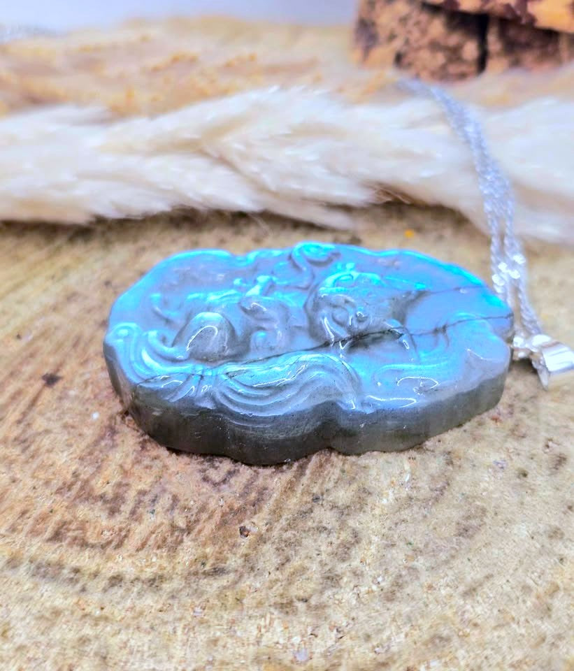 A carved stone pendant with a silver-toned chain, featuring a raised animal motif and a bright blue-green iridescent sheen, displayed on a textured wood surface.