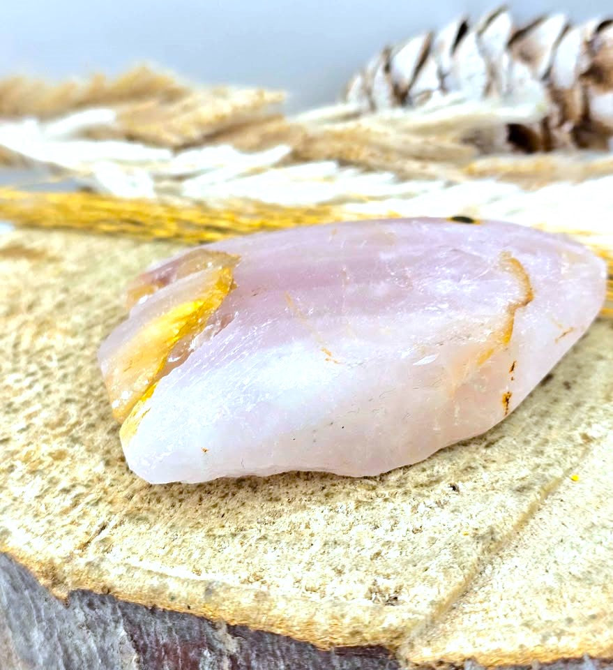 A polished pale pink and white crystal stone with a gold fissure, resting on a textured wood slice with dried grass and a pinecone in the background.