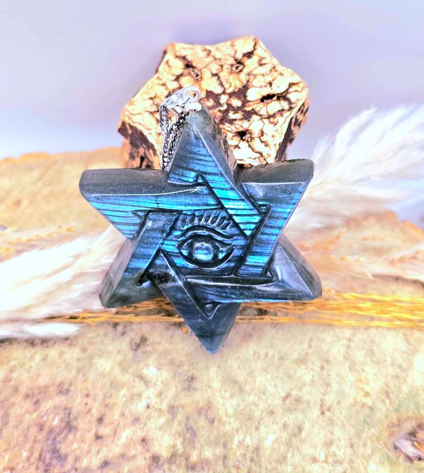 A dark metallic star-shaped pendant with a blue iridescent sheen and an eye motif in the center, displayed on a textured cork piece with dried grass against a soft lavender background.