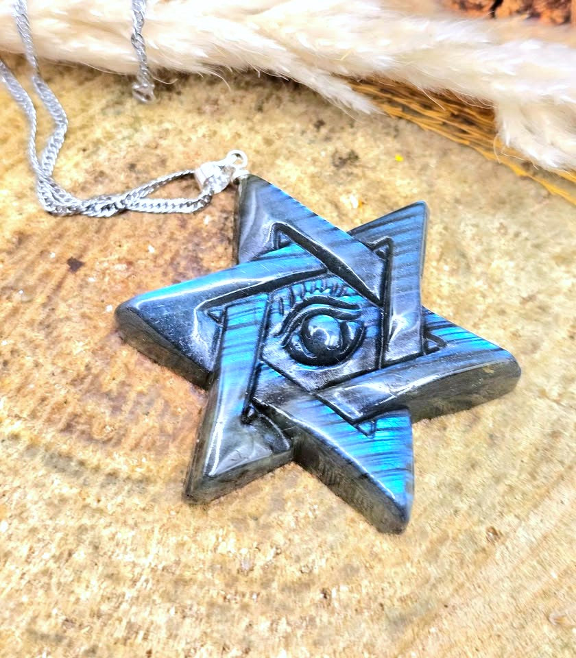 A star-shaped pendant necklace with a silver-toned chain, featuring layered geometric star shapes in black and iridescent blue-green, with an eye motif at the center, displayed on a textured wood surface.
