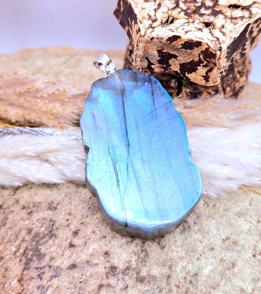 A polished blue labradorite pendant with natural inclusions, resting on a textured stone surface with a white feather and wood background.