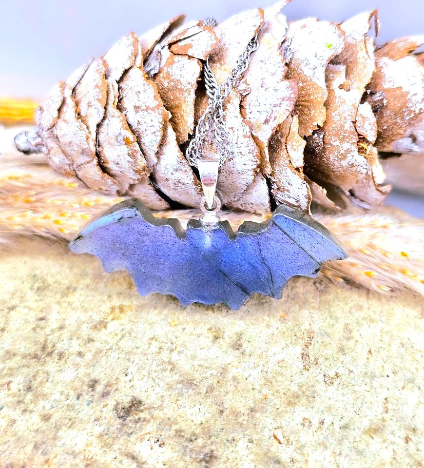 A silver bat-shaped pendant necklace with a blue iridescent stone inlay, displayed on a pinecone and textured wood against a light background.