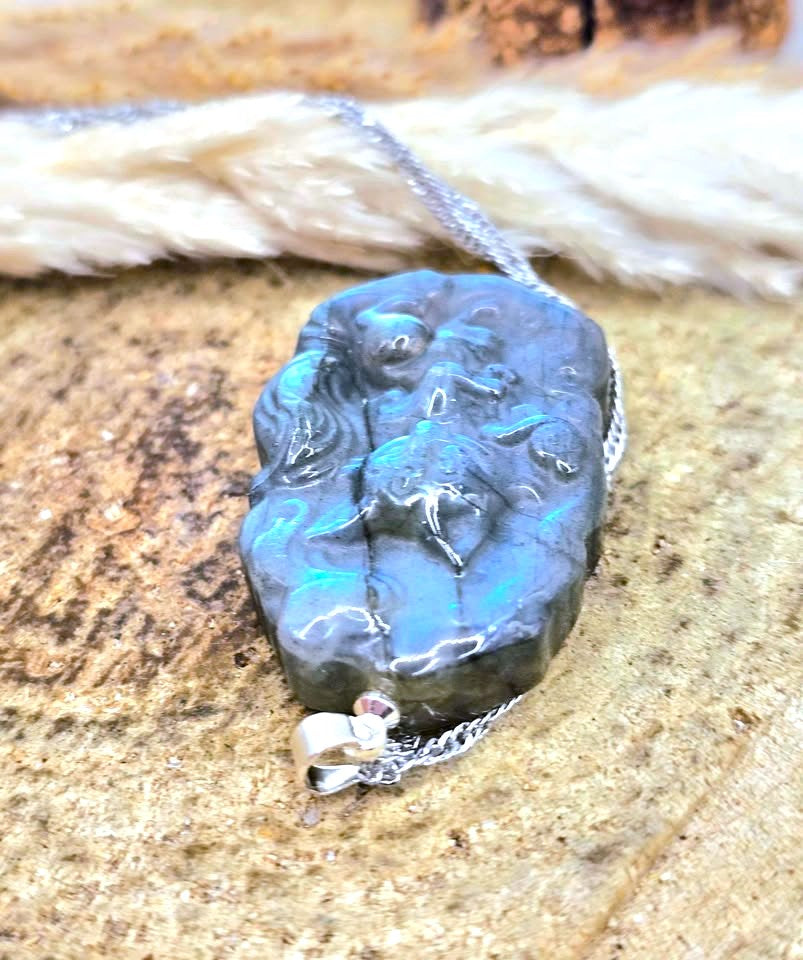A raw, polished dark gray stone pendant with vibrant turquoise blue iridescence, suspended on a silver chain with a small silver bail, resting on a textured tan stone surface.