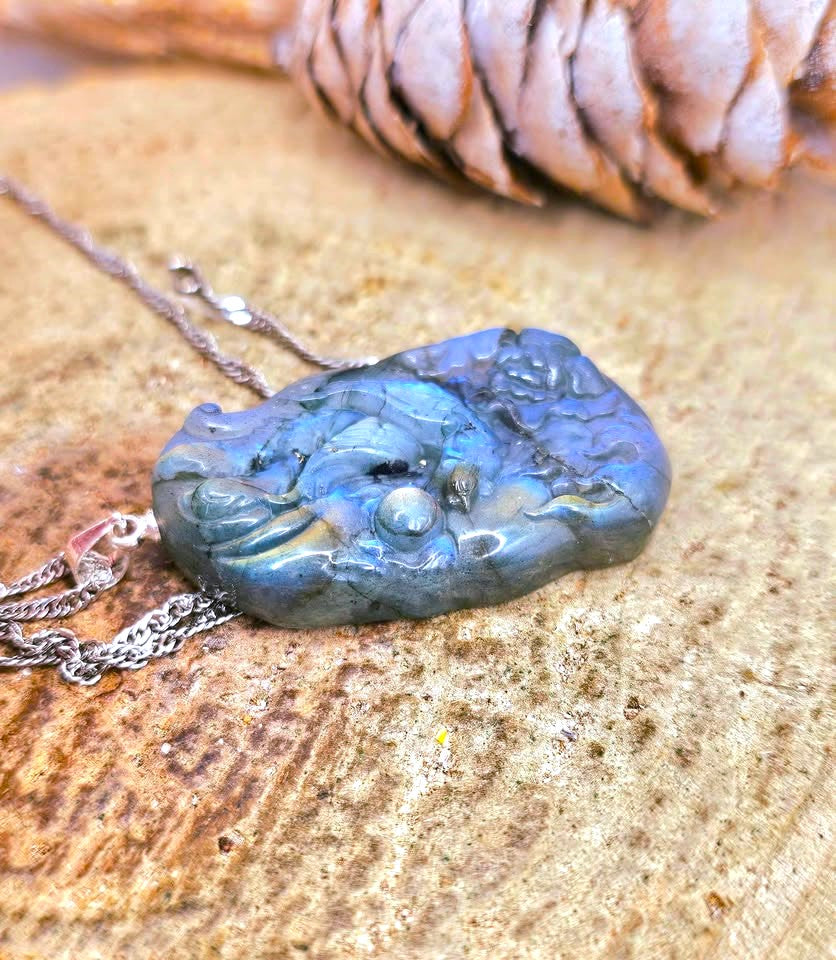 A carved labradorite pendant necklace with a silver-toned chain, resting on a textured wood surface with a pinecone in the background.