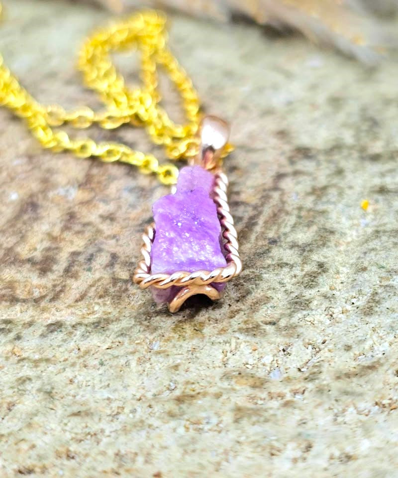A close-up of a necklace with a rough purple stone pendant in a twisted metal setting, hanging from a gold-tone chain on a textured stone surface.