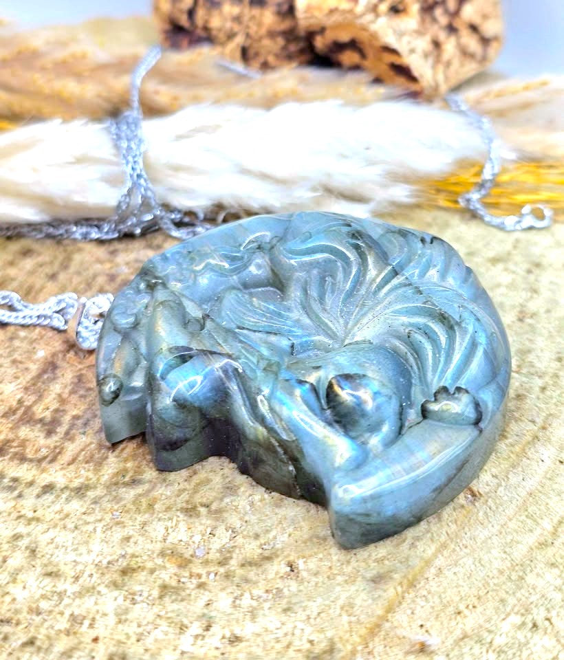 A carved oval pendant necklace made of polished labradorite with a silver-toned chain, resting on a textured stone surface with dried grass and bark in the background.