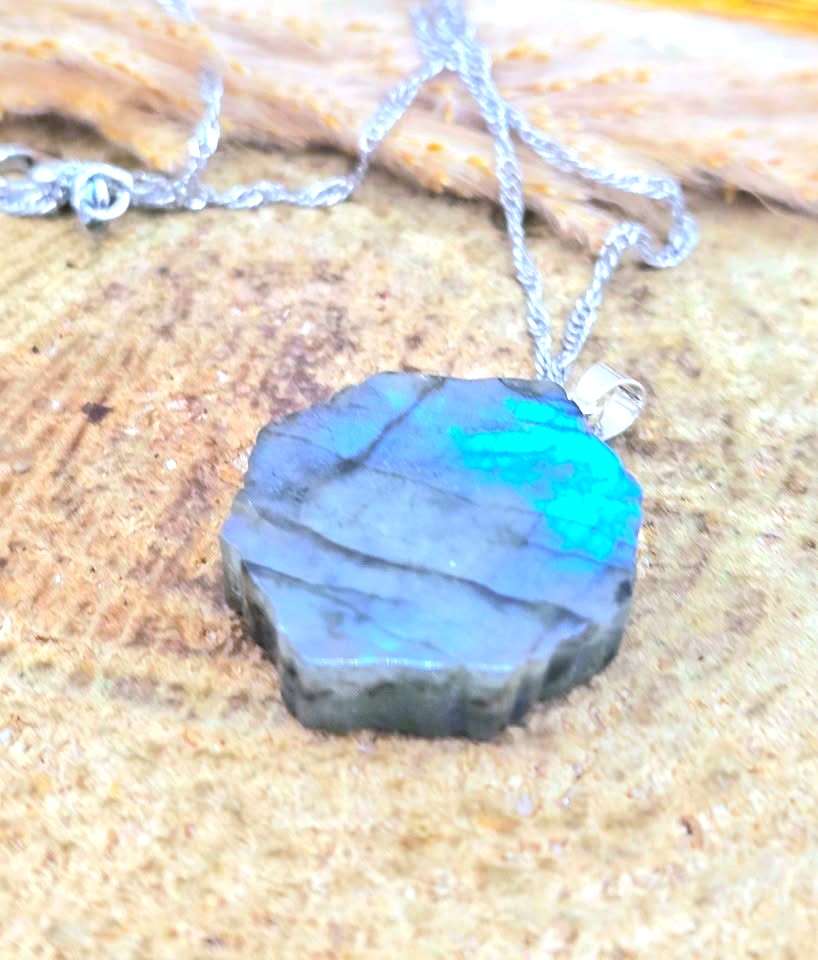 A silver chain necklace with a polished, irregular-shaped labradorite pendant showing vibrant blue and green flashes, resting on a textured wood surface.