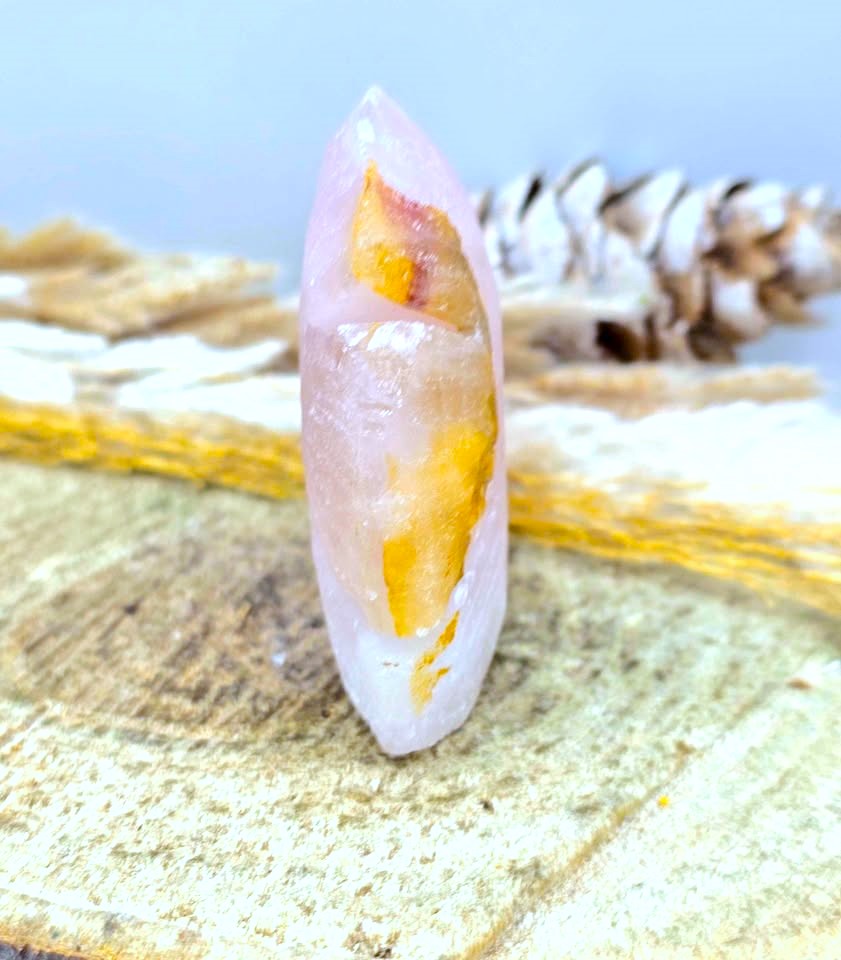A pink rose quartz crystal point with prominent golden-yellow inclusions, displayed on a textured wooden slice with a pinecone in the blurred background.