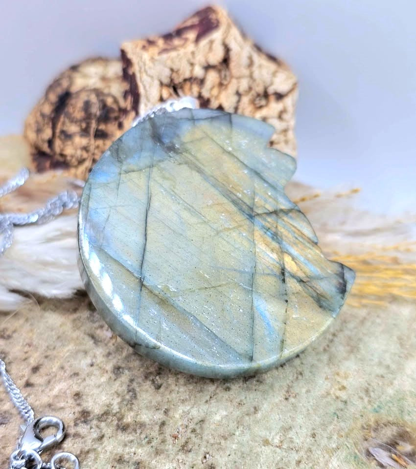 A polished round labradorite pendant necklace with a silver clasp, displayed on a textured stone surface with a cork piece in the background.