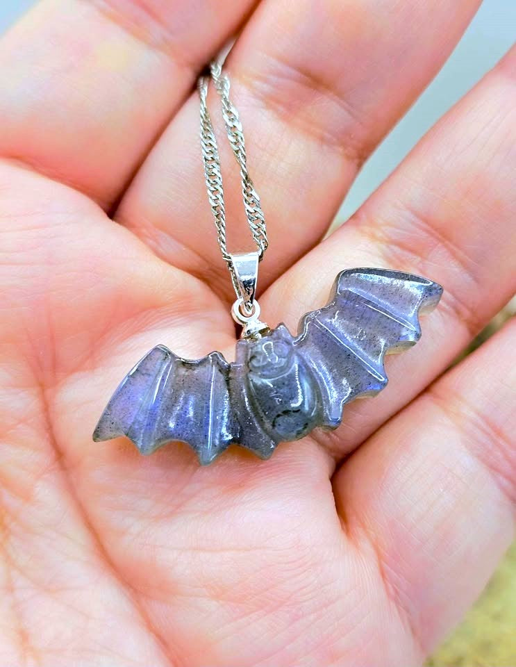 A bat-shaped pendant necklace made of polished gray stone with a silver chain, held in a person's hand.