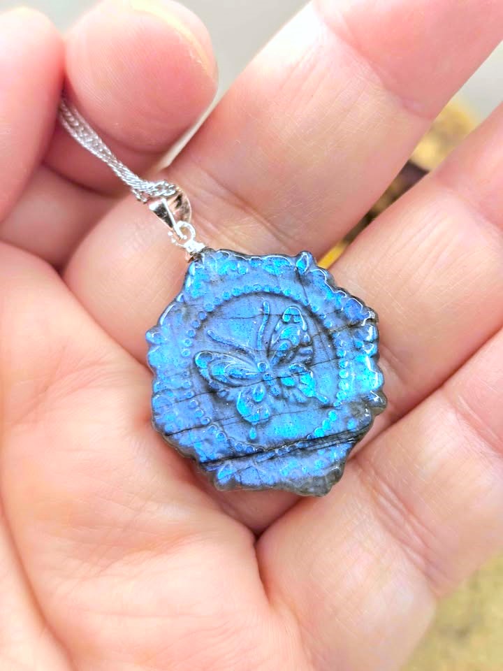 A close-up of a hand holding a blue iridescent wax seal pendant necklace with a butterfly design, attached to a silver chain.