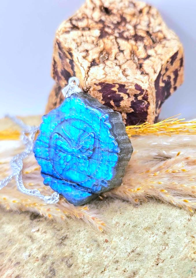 A blue and black textured pendant with a carved emblem, resting on a piece of cork bark and dried grass against a light background.