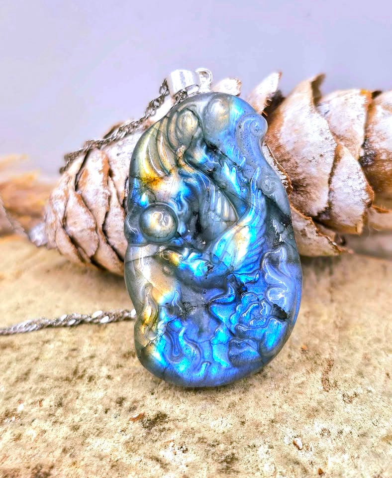 A carved labradorite pendant necklace featuring a mermaid design with iridescent blue, green, and gold flashes, resting on a pinecone and textured stone surface.