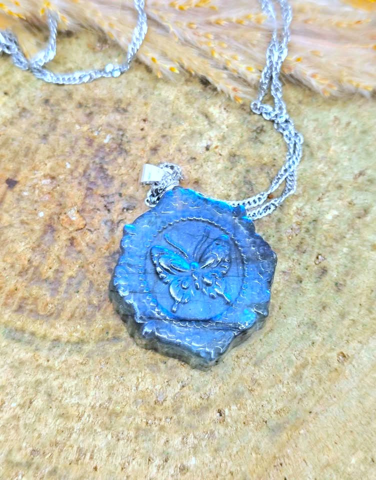 A silver chain necklace with a textured, irregularly shaped blue pendant featuring an embossed butterfly design, displayed on a textured tan stone surface.