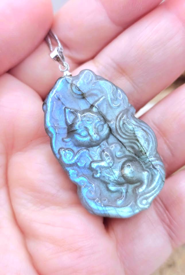 A close-up of a hand holding a carved pendant with a silver bail. The pendant has an iridescent blue-green finish with raised animal and wave motifs.