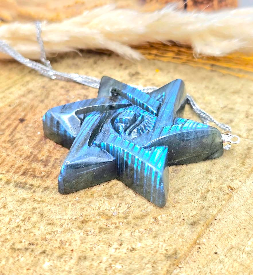 A five-pointed star pendant necklace with a dark metallic finish and bright blue painted accents, resting on a textured tan surface with dried grass in the background.