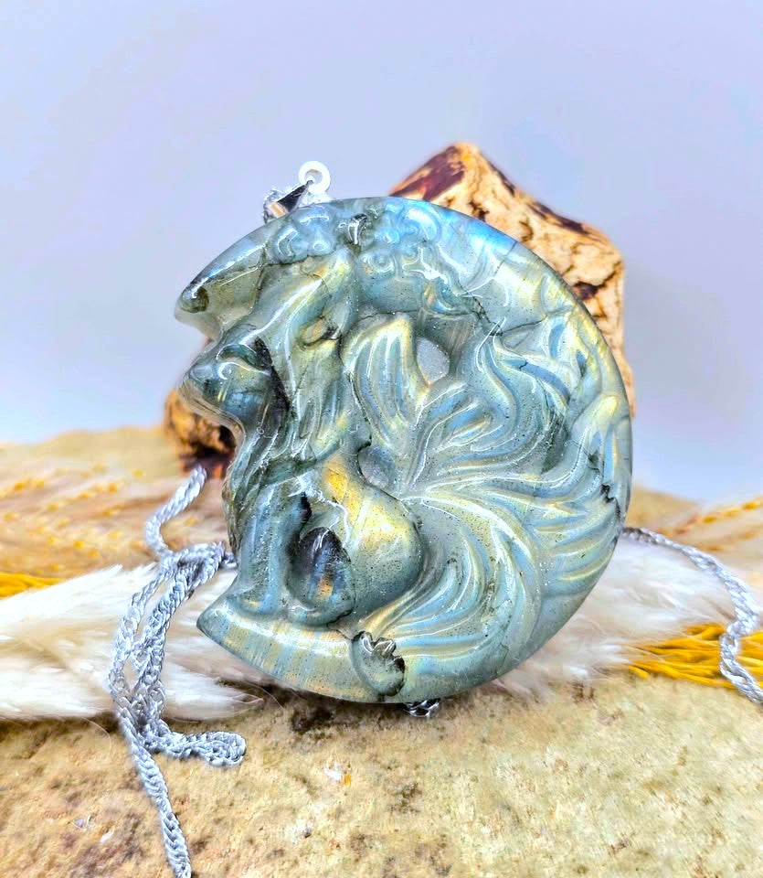 A carved crescent moon pendant featuring a stylized face with flowing hair, shown resting on a textured surface with a silver chain.