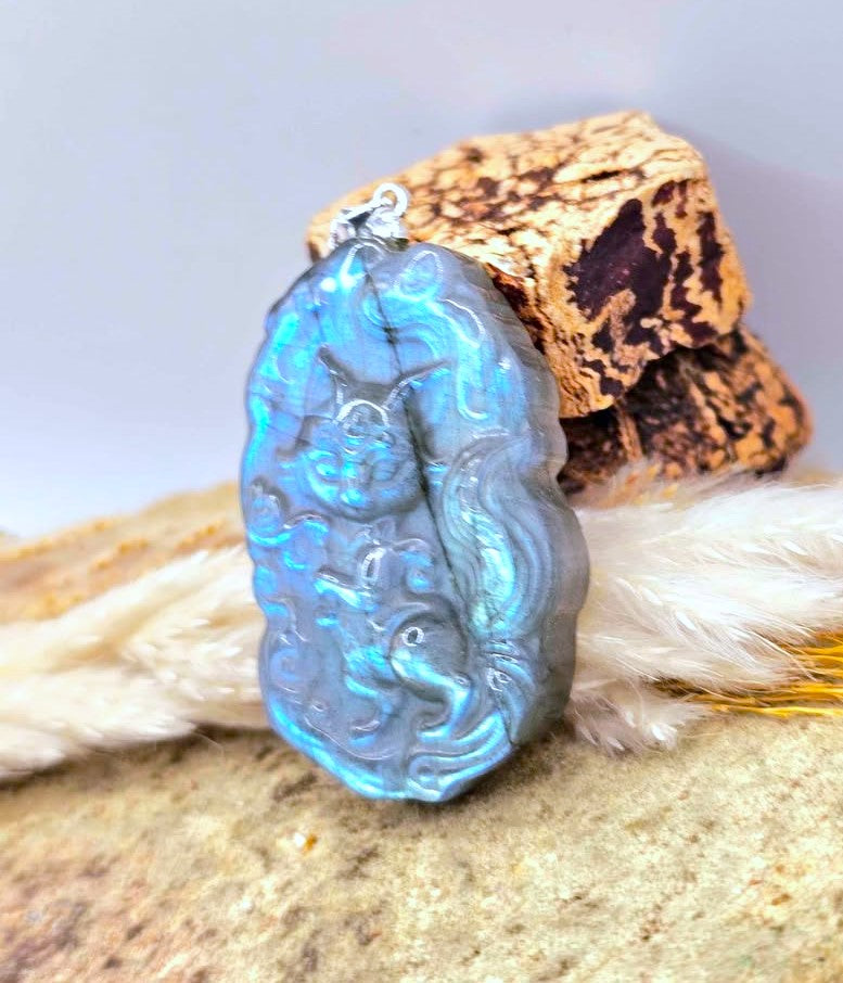 A carved blue labradorite pendant featuring a stylized figure, displayed on a textured stone surface with a piece of wood and white feathers in the background.