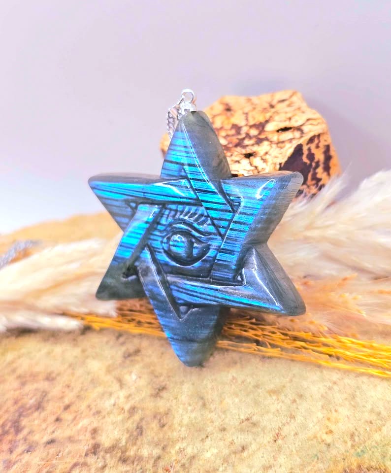 A blue and black striped star-shaped pendant with an eye design in the center, resting on dried grass and a cork against a soft lavender background.