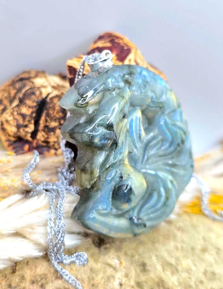 A carved oval pendant necklace made of iridescent blue-green stone with a silver bail and chain, displayed on a textured beige surface with dried botanicals in the background.
