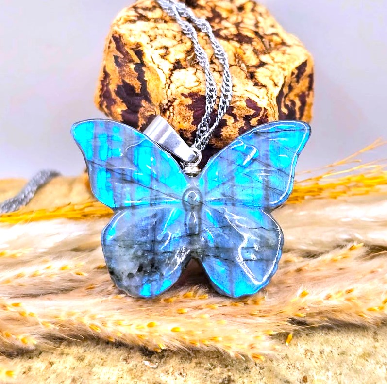 A butterfly-shaped pendant necklace made of iridescent blue labradorite stone with a silver chain, displayed on a textured cork block and dried wheat stalks.