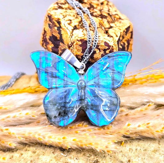 A butterfly-shaped pendant necklace made of iridescent blue labradorite stone with a silver chain, displayed on a textured cork block and dried wheat stalks.