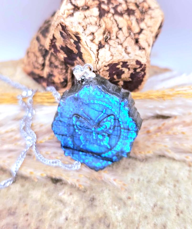 A hexagonal blue labradorite gemstone pendant with a butterfly motif, hanging from a silver chain, styled on a textured beige surface with a cork piece in the background.