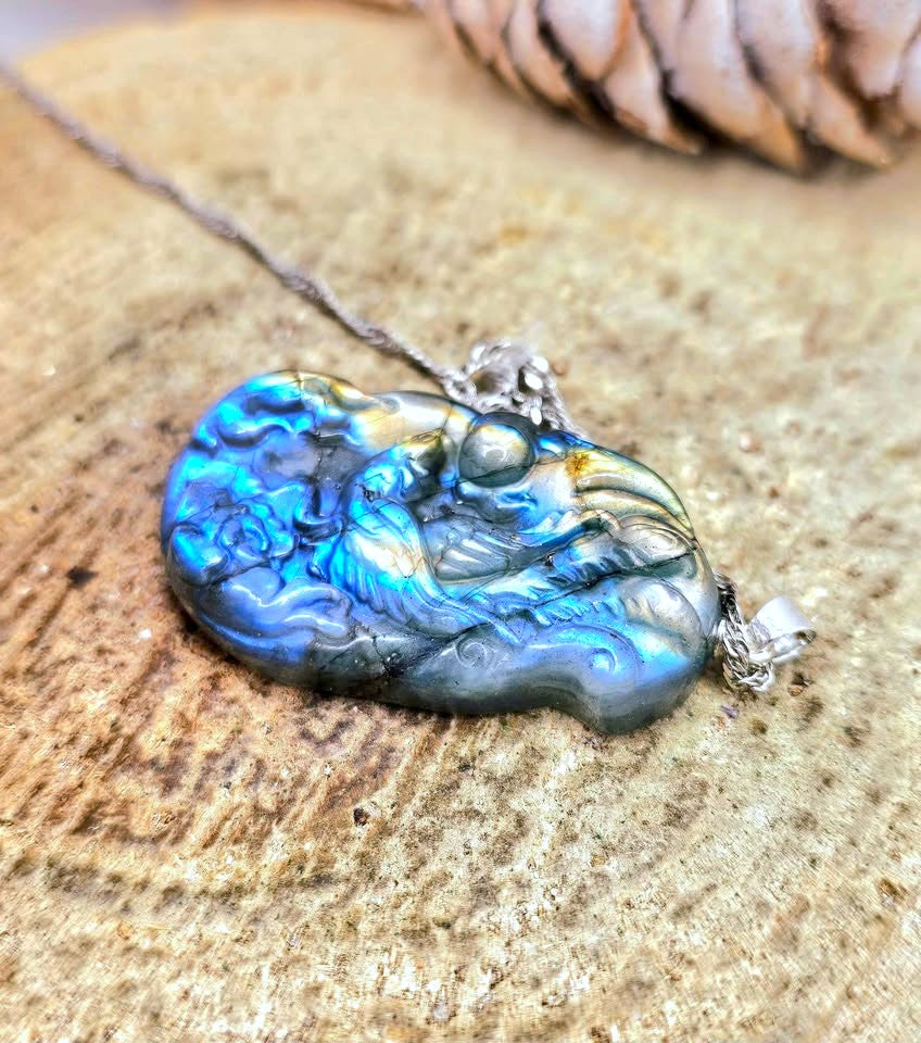 A close-up of a carved pendant necklace with a bird design, featuring a vibrant blue and green iridescent finish, resting on a textured stone surface.