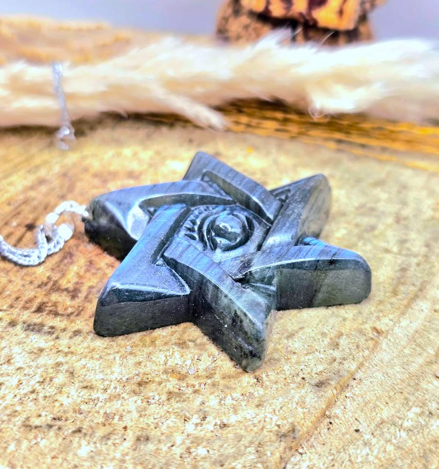 A carved black stone pentacle pendant with an eye of providence symbol, resting on a textured wooden surface with a white cord.