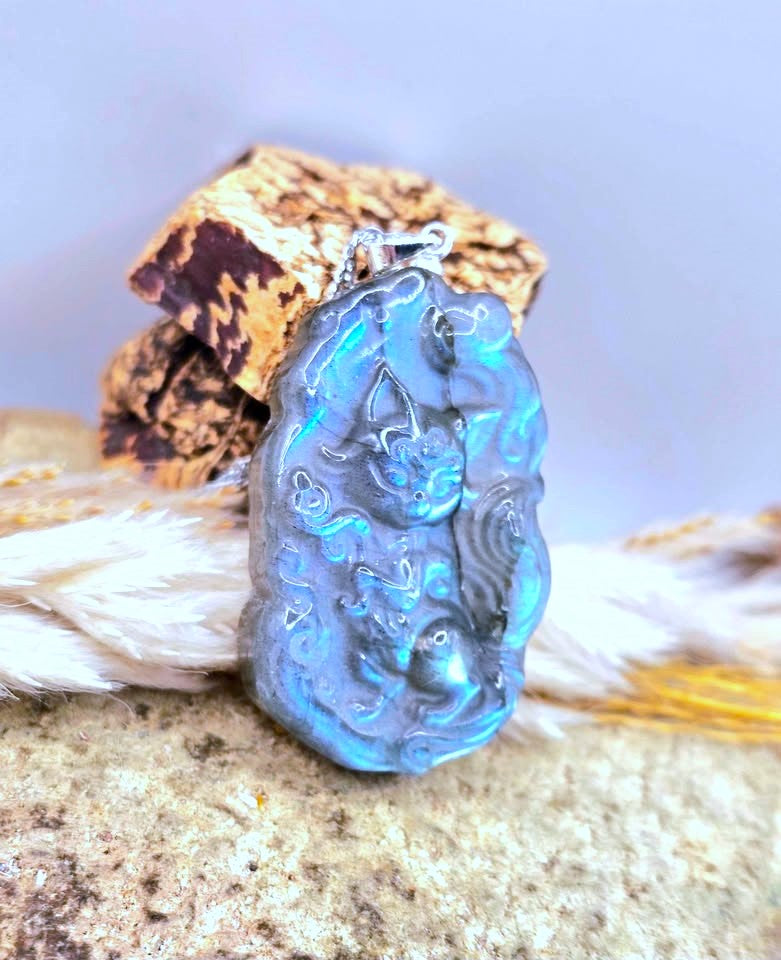 A carved blue-gray stone pendant with a silver bail, displayed on a textured rock with dried grass and bark against a soft light background.
