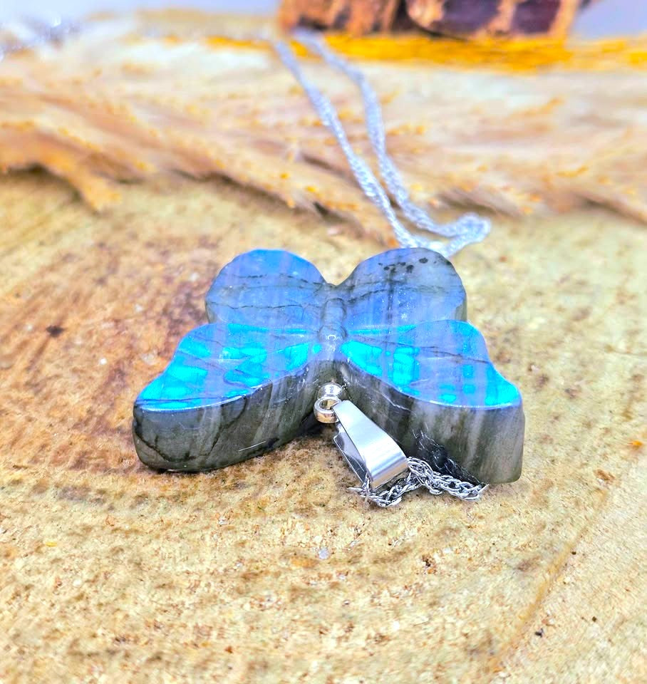 A butterfly-shaped pendant necklace made from dark stone with vibrant blue and green iridescent inlay, hanging from a silver-toned chain on a textured sandy surface.