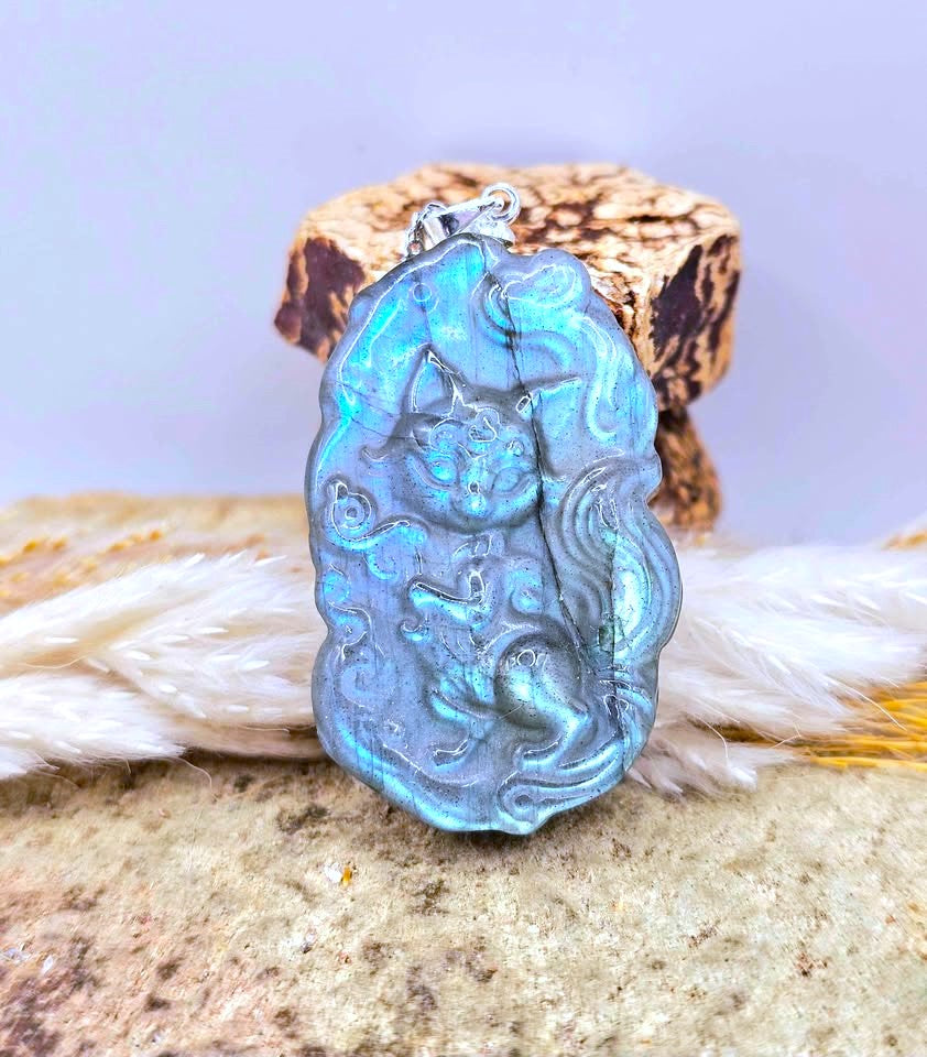 A carved pendant featuring a fox or cat-like figure with an iridescent blue-green finish, suspended from a silver loop and displayed against a soft lavender background with decorative white feathers and a textured stone surface.