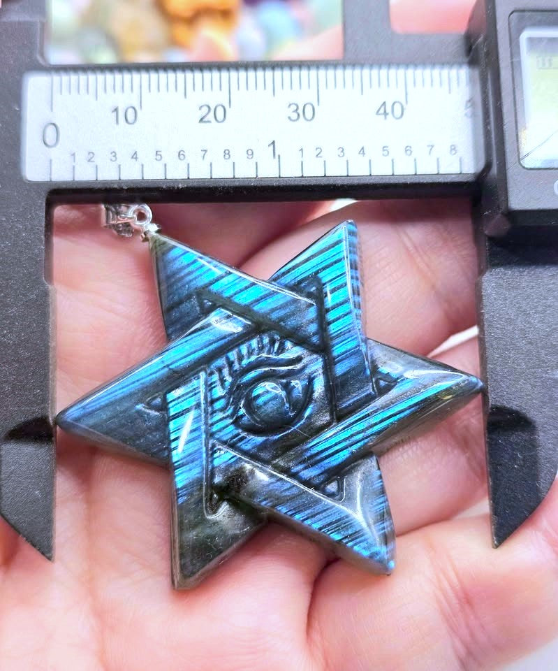 A close-up of a metallic star-shaped pendant held in a hand under a digital caliper. The pendant has a glossy blue and black iridescent finish with a central eye motif.