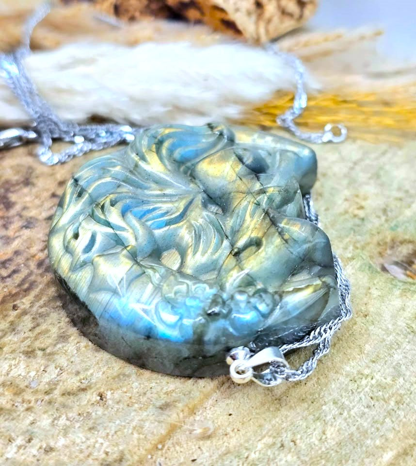 A carved labradorite pendant necklace with a silver chain, resting on a textured stone surface. The pendant features natural blue and gold labradorescence, and the silver chain is partially visible.