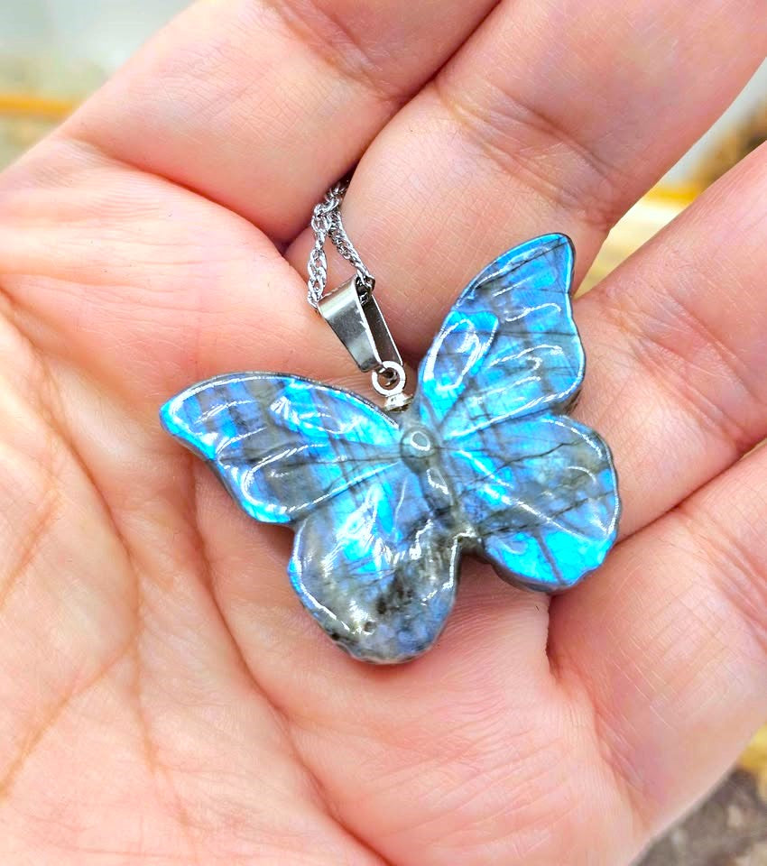 A butterfly-shaped pendant made from iridescent blue labradorite stone, held in a person's hand. The pendant is attached to a silver chain.