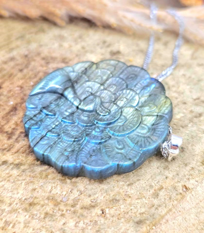 A round, hand-carved labradorite pendant necklace with a silver chain, resting on a textured sandy surface.