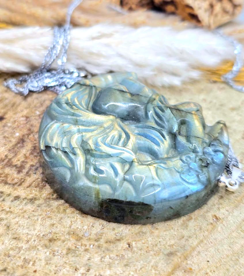A carved labradorite pendant necklace featuring a stylized figure, resting on a textured stone surface with a silver chain.