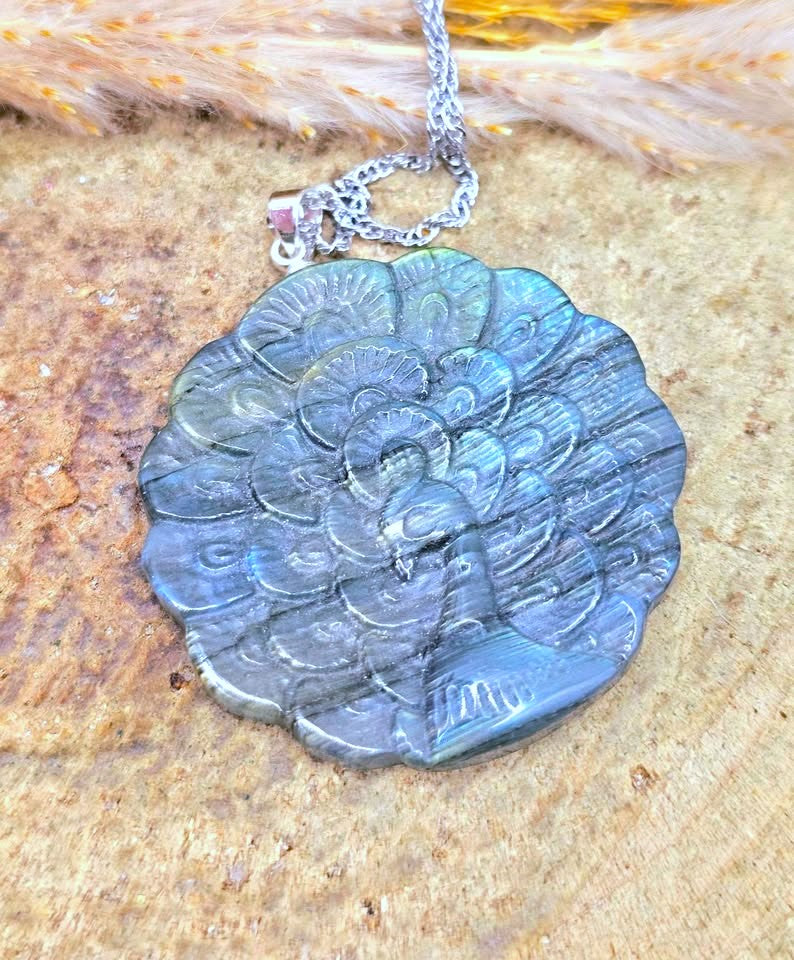 A silver chain necklace with a round carved pendant featuring a peacock design, displayed on a textured wood surface with dried grass.